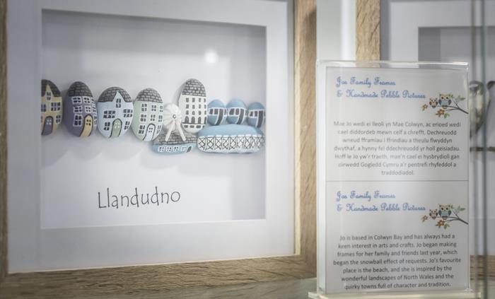 Handmade pebble art frame showing a Llandudno seaside scene alongside information about a local artist from North Wales.